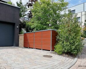 Garage mit Rollladen, gepflasterte Einfahrt, Holzverkleidung vor Büschen und Bäumen, modernes Gebäude im Hintergrund.