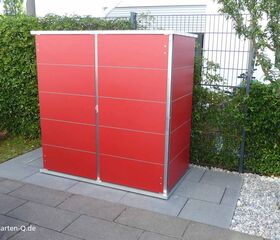 . Ein rotes Gartenhaus mit Metallrahmen auf einer gepflasterten Fläche, umgeben von Pflanzen und Bäumen.