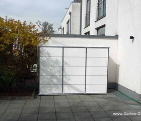 . Ein weißer Schrank steht auf einer Terrasse neben einem Gebäude und Büschen.
