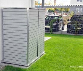. Ein moderner Gartenschrank aus Metall steht neben einem Haus auf einer grünen Rasenfläche. Im Hintergrund parken Autos.