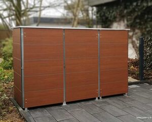 . Holzverkleideter Mülltonnenunterstand im Garten auf gepflastertem Boden vor Bäumen und Sträuchern.