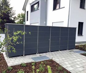 . Ein moderner, grauer Gartenschrank steht auf einer gepflasterten Fläche neben einem Haus mit weißem Anstrich.
