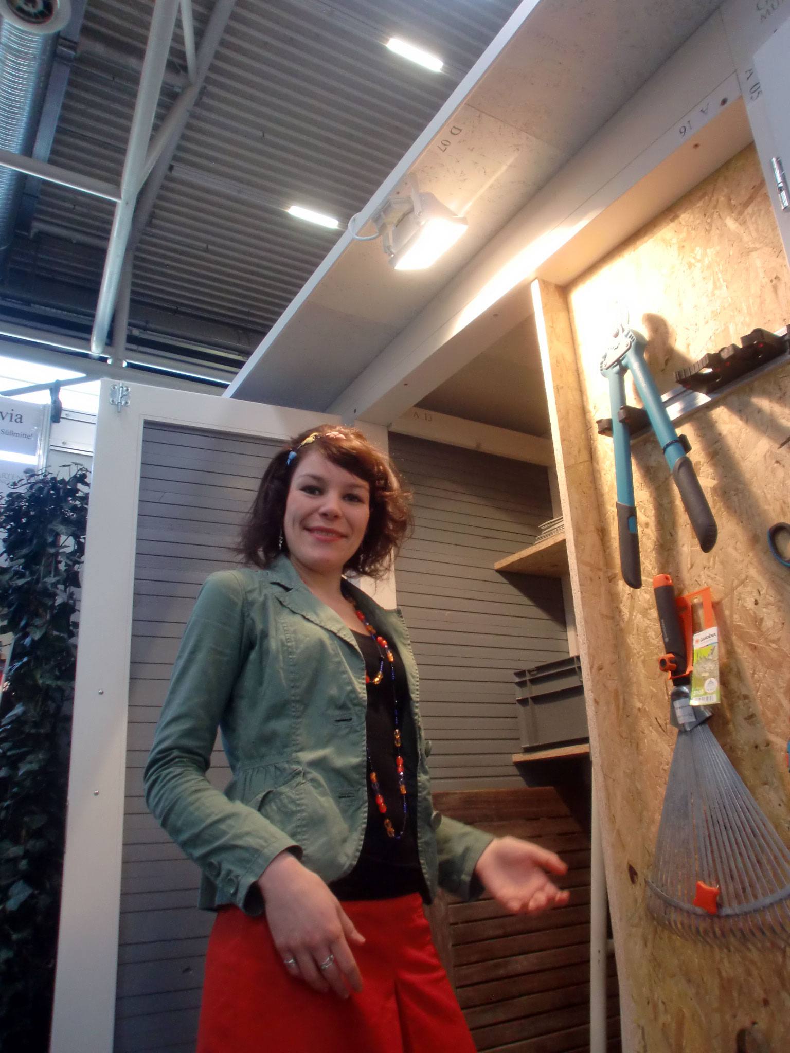 . Eine Frau steht in einem Baumarkt neben Werkzeugen, darunter eine Astschere und ein Rechen.